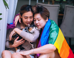 Happy lesbian couple with rainbow flag enjoy playing with their pet cat. Togetherness concept.
