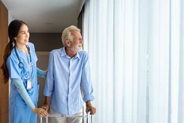 Hospice nurse is supporting Caucasian man to use walker while looking out the window in pension retirement center for home care rehabilitation and post treatment recovery process - obrazy, fototapety, plakaty