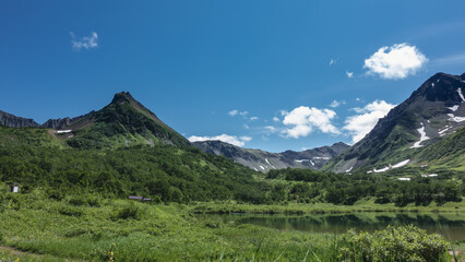 A calm lake on an alpine meadow. Lush green vegetation on the banks. A picturesque mountain range against a background of blue sky and clouds. Kamchatka. Vachkazhets. Lake Tahkoloch
