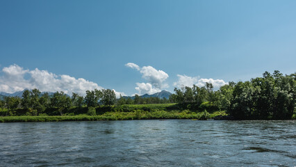 A calm blue river. There is lush green vegetation on the shore. Mountains on the background of azure sky and clouds. Kamchatka. River Bystraya