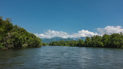 Fototapeta na wymiar A calm blue river under an azure sky. There are thickets of green trees on the banks. A mountain range in the clouds. Kamchatka. River Bystraya