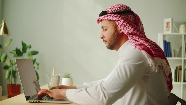 Middle eastern man using laptop. Home office. Male person texting at computer keyboard in living room. Wearing traditional Islamic clothes. Communicating with family and friends online.