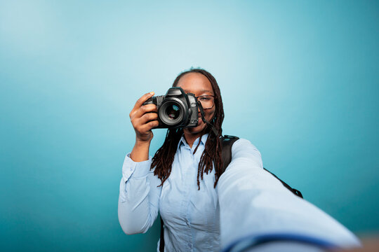Creative amateur photographer taking pictures with modern camera for social media. Young adult african american woman pointing DSLR device to camera while taking a photo on blue background.