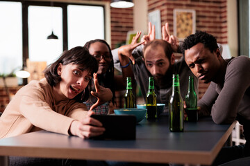 Goofy happy friends taking selfie group photo with modern smartphone while acting childish. Positive young adult people sitting at home in living room with mobile cellphone while taking picture.