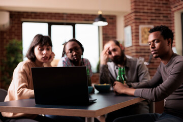 Multiethnic friends sitting at table while watching video content on laptop. Diverse group of people at home in living room enjoying movies together on modern portable computer while having snacks.