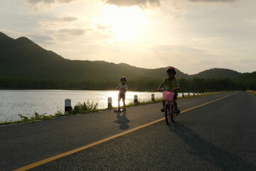 Cute little girl riding a bicycle and her sister riding a scooter on a lake road at sunset. Happy sisters doing outdoor activities together. Healthy Summer Activities for Kids.