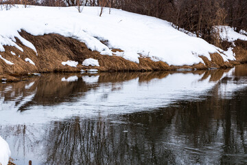 forest river with fast flow and small waves flows between snowy shores with trees and bushes late winter and early spring
