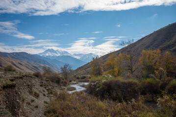 Mountain valley with a small river and a country road. A mountain range with snow-capped peaks in a blue haze in the distance. Bright autumn sky with beautiful clouds and yellow trees.