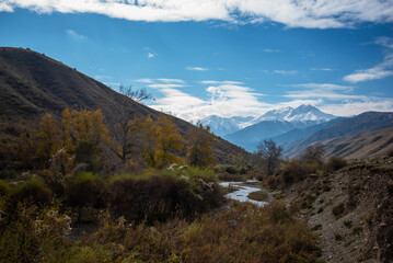 Mountain valley with a small river and a country road. A mountain range with snow-capped peaks in a blue haze in the distance. Bright autumn sky with beautiful clouds and yellow trees.
