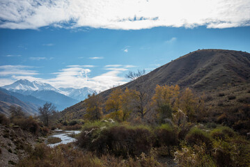 Obraz premium Selective focus. Mountain valley with a small river and a country road. A mountain range with snow-capped peaks in a blue haze in the distance. Autumn sky with beautiful clouds and yellow trees.