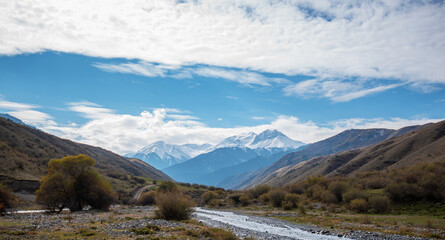 Mountain valley with a small river and a country road. A mountain range with snow-capped peaks in a blue haze in the distance. Bright autumn sky with beautiful clouds and yellow trees.