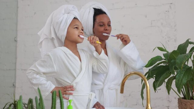 Young mother, daughter are brushing their teeth and having good morning together at home spbi. 4k Close view of american african woman, cute girl brush teeth and look with smiles, talk and stand in