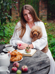 middle aged caucasian woman in white dress enjoying morning coffee in apple orchard sitting at wooden table with her pet poodle