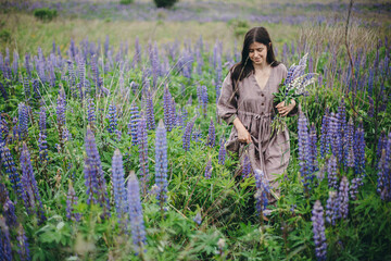 Cottagecore aesthetics. Stylish woman in rustic dress holding lupine bouquet in meadow. Young female in linen dress gathering wildflowers in atmospheric summer countryside, rural slow life