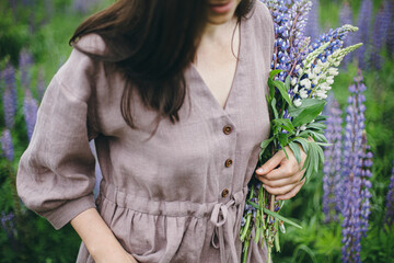 Close up of woman in rustic dress holding lupine bouquet in meadow. Cottagecore aesthetics. Young...