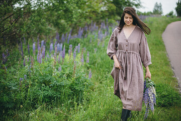 Stylish woman in rustic dress walking with lupine bouquet in summer countryside. Cottagecore aesthetics. Young female in linen dress holding wildflowers after rain on rural road, slow life