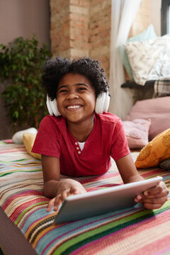 African happy boy in wireless headphones lying on sofa using digital tablet for funny games