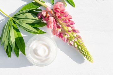 A white cosmetic jar with face natural cream on a white background next to pink lupine and green leaves. Natural organic skin care cosmetics