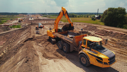 A yellow digger filling up a tipper truck with sand building a new long highway in the middle of a countryside. High quality photo - obrazy, fototapety, plakaty