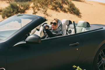 Fototapeta na wymiar Glamorous blonde in a silk scarf and sunglasses in the driver's seat of a convertible convertible on a hot desert background. Fashion, style, glamour