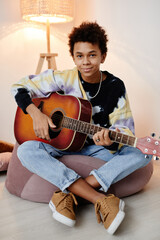 Vertical full length portrait of black teenage boy playing guitar and smiling at camera while...