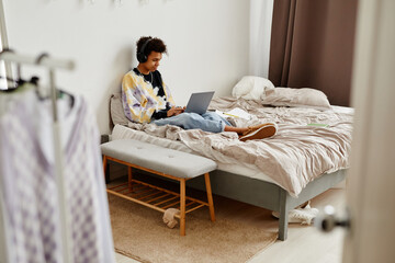 Wide angle view at black teenage boy with laptop sitting on bed in messy room, copy space