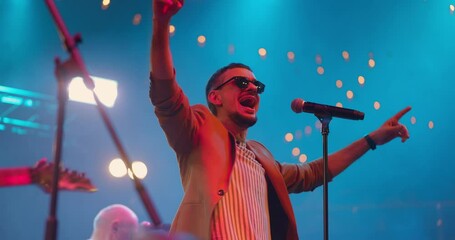 Attractive young male singer in glasses performing with band at live music event. Portrait handsome musician singing song in microphone on pop concert. Stage light, smoke