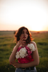 Fototapeta na wymiar Stylish girl with long wavy hair holds a bouquet of peonies in the field at sunset.