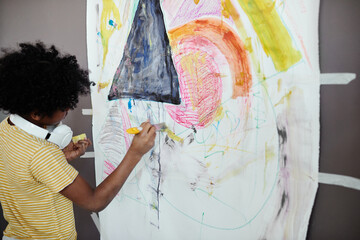 African little boy using yellow crayon to draw a picture on big paper hanging on the wall