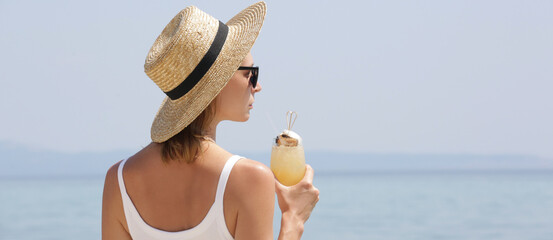 Young woman with straw hat holding a glass of cocktail and looking at the horizone over the sea....