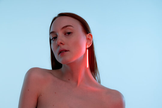 Portrait of tender young girl with neon colored accessories, earrings posing, looking at camera isolated over blue background