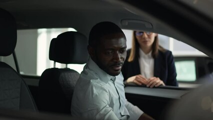 Young businessman is sitting inside car and talking to salesperson in dealership office spbas. 4k Close view of bearded African man examines new car and looks with smile, consults with female manager