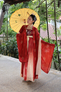 Young attractive Asian woman wearing tadeonal Chinese red hanfu long skirt dress costume scarf hairpin earing decorated umbrella outdoor green garden patio