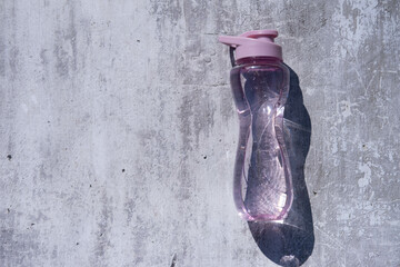 A flat shot of a plastic, reusable bottle of isotonic drink on a gray concrete background with hard shadows from the sun, a space for copying on the left side