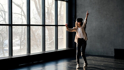 Fototapeta na wymiar A teenage girl in virtual reality glasses stands in a gray room near a large window.