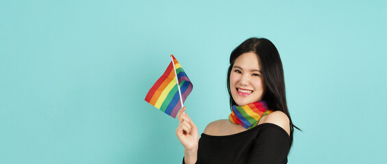 LGBTQ woman holding pride flag standing against a blue green background. Asian LGBTQ woman with rainbow scarf on neck. look smart bright and energetic cheerful. LGBTQ diversity pride concept.