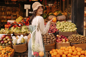 Beautiful young caucasian woman smiling turning to camera standing near fruit stand. Brunette wears casual clothes and shopping bag with groceries. Healthy food concepts