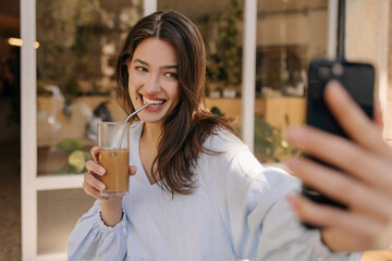 Smiling young caucasian woman drinking cocktail takes selfie on phone sitting outdoors. Brunette wears casual dress in summer. good morning concept