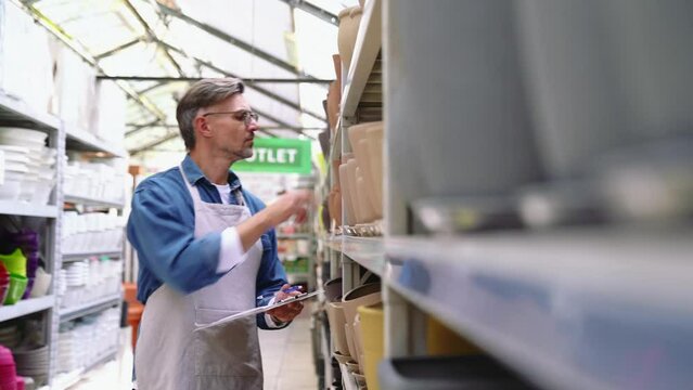 Serious grey haired florist man checks and writes about flower pots indoors