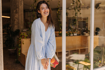 Smiling young caucasian woman looking at camera, holding hands together standing indoors during daytime. Brunette wears blue sundress in summer. Lifestyle, different emotions, leisure concept