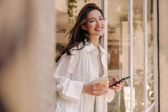 Beautiful young caucasian woman smiling looking at camera, holding phone and glass standing in doorway. Brunette wears white shirt warm weather. Mood, lifestyle, concept.