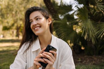 Cute young caucasian girl smiling looking at camera, holding smartphone in hands being outdoors. Brunette wears casual white shirt. Concept of use