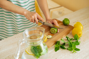 Hands of a woman preparing homemade mint, lemon, lime lemonade and a glass jug of lemonade. Cutting a lime on a wooden board for homemade lemonade. © Nicole's