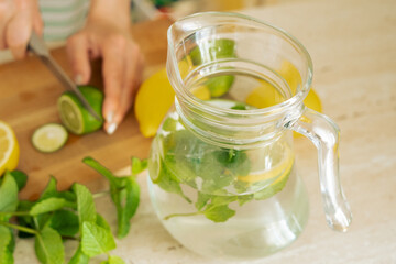 Hands of a woman preparing homemade mint, lemon, lime lemonade and a glass jug of lemonade. Cutting a lime on a wooden board for homemade lemonade.