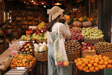 Fair-skinned young girl holds orange standing with her back to camera in fruit street shop. Brunette wears hat, blouse, pants and string bag. Concept shopping