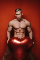Naked fitness man with inflatable big heart at red background. Shirtless male model with six pack...