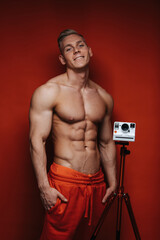 Shirtless young and handsome male model in red pants near tripod with camera at red background....