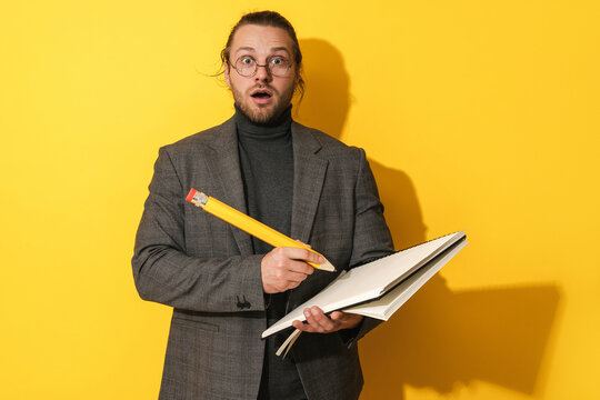 Funny man wearing glasses holding big pencil and notebook on yellow background