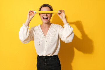 Excited woman covering her eyes with big pencil on yellow background