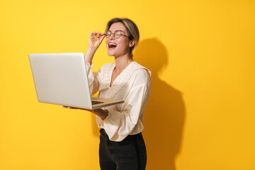 Cheerful woman wearing eyeglasses is using laptop computer on yellow background